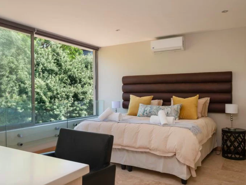 A bright and modern bedroom featuring large sliding windows with a glass balustrade, stone flooring, and a wall-mounted air conditioning unit.