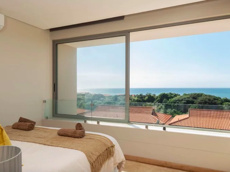 Modern bedroom featuring a large sliding window with expansive ocean and coastal views, complemented by a sleek glass balustrade and integrated air conditioning.