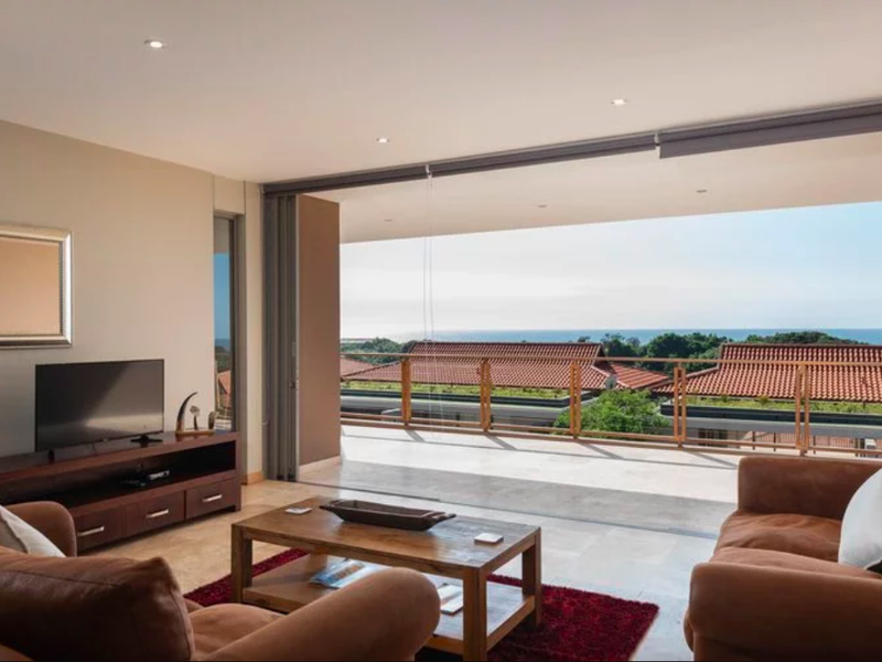 Spacious living room featuring large sliding glass doors that open onto a balcony with scenic coastal views and tiled flooring throughout.