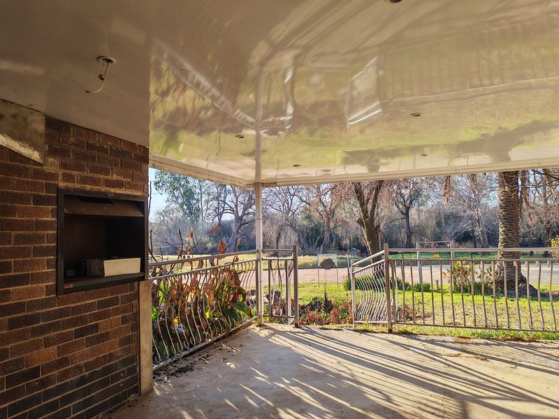 Covered patio area featuring a built-in braai, concrete flooring, and metal railings, overlooking a garden setting. Requires minor maintenance to railings and electrical fixtures.