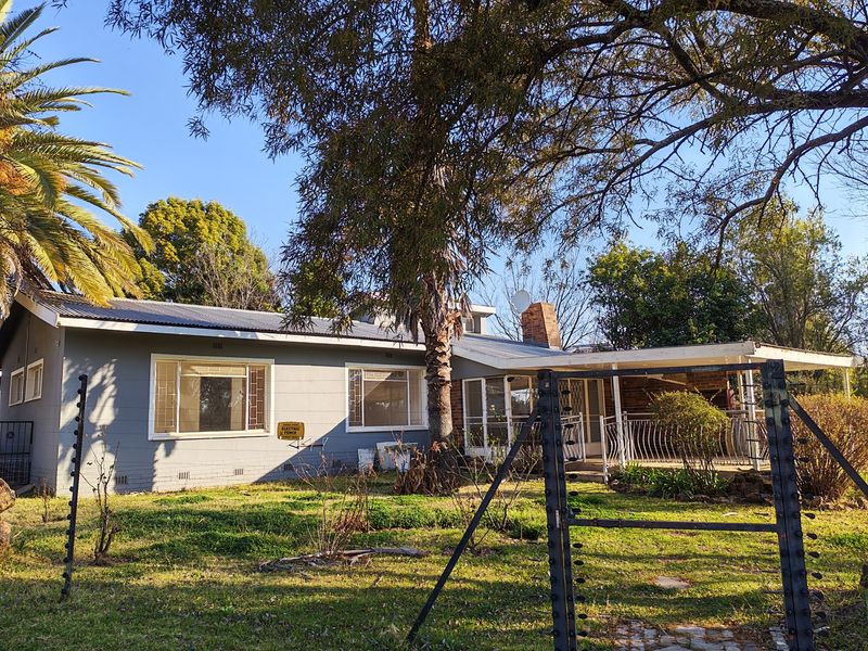 Front view of a single-story house featuring a corrugated metal roof, brick chimney, covered patio area, and perimeter electric fencing.
