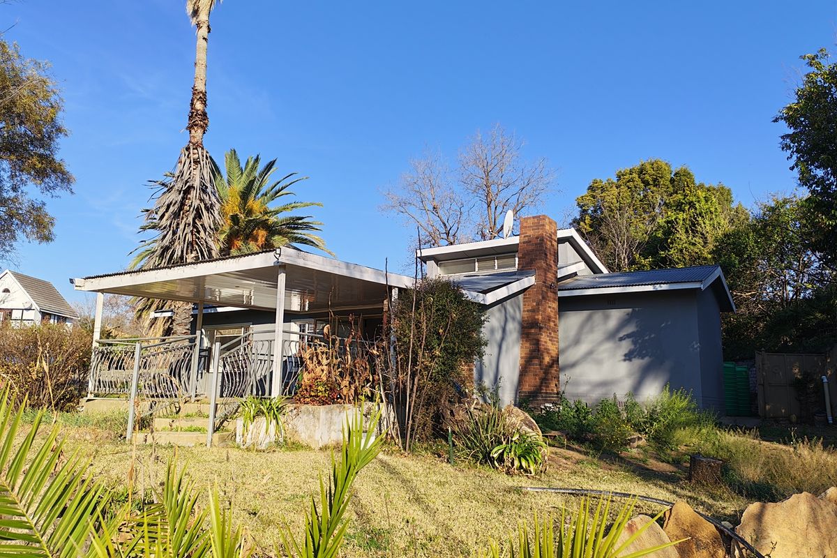 Rear view of the house featuring a covered patio, brick chimney, and metal roofing, set within a garden with established trees and landscaping.