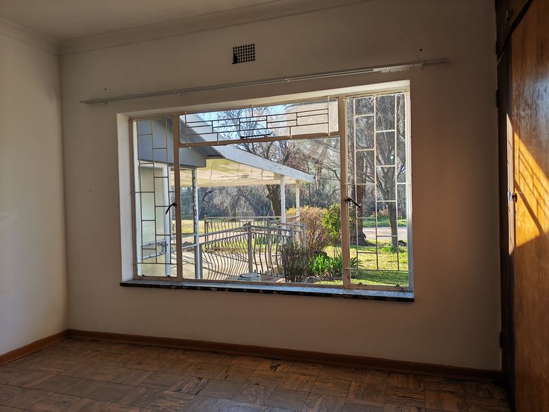 An empty living room featuring parquet flooring, a large window with security bars, and a built-in cupboard, offering a view of the garden.