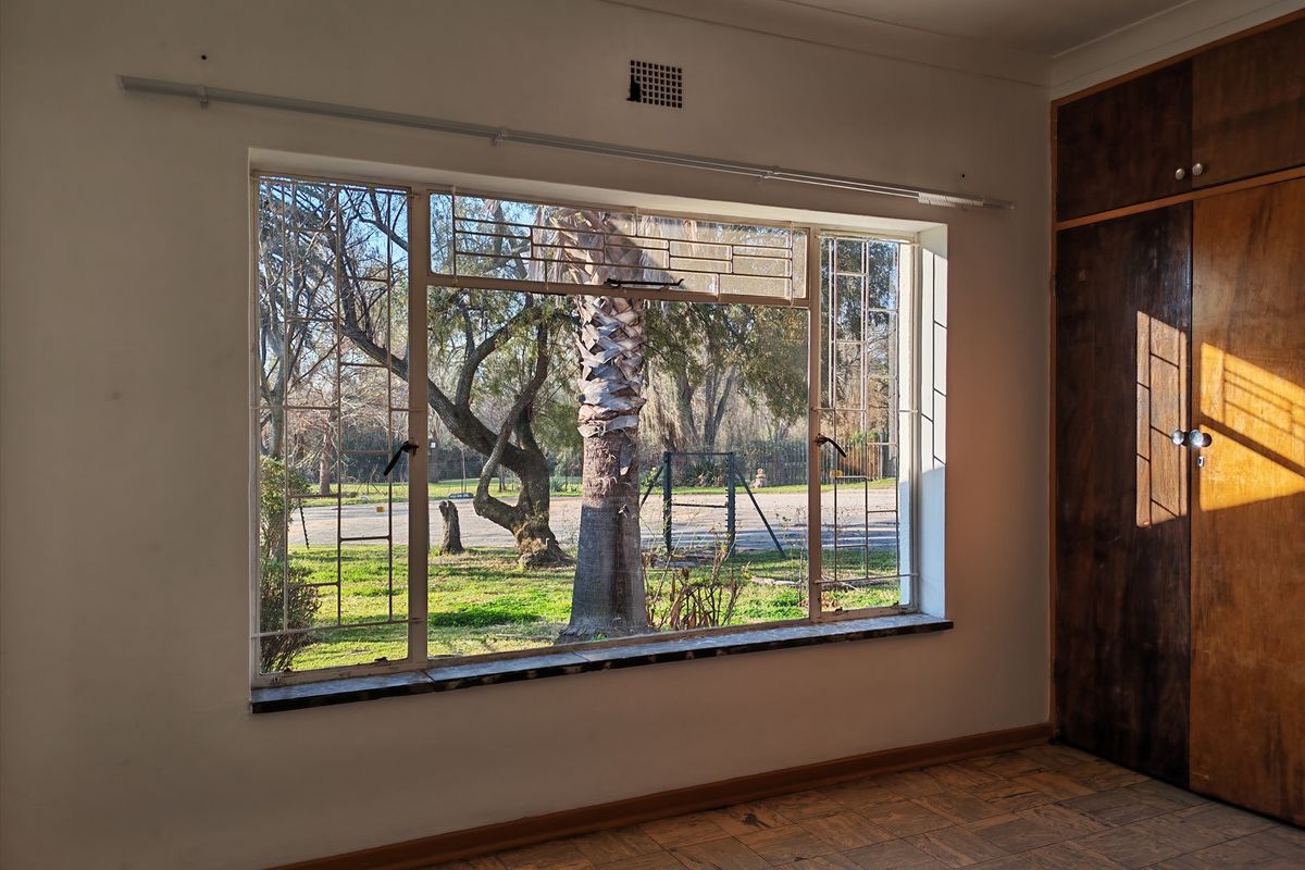 An empty bedroom featuring original parquet flooring, built-in wooden cupboards, and a large steel-framed window with security bars overlooking the garden.
