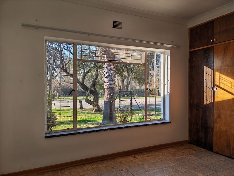 An empty bedroom featuring original parquet flooring, built-in wooden cupboards, and a large steel-framed window with security bars overlooking the garden.