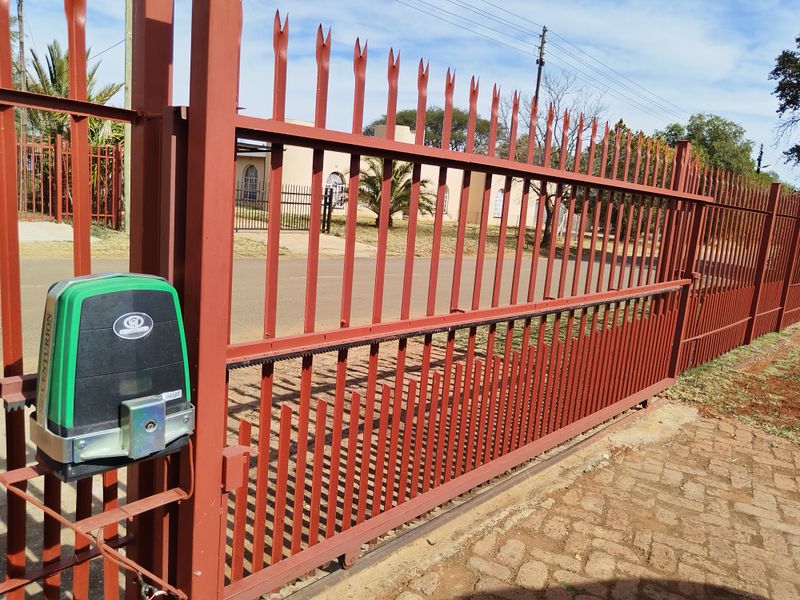 A secure sliding gate featuring an automated electric motor and durable steel palisade fencing, providing access to the property's paved driveway.