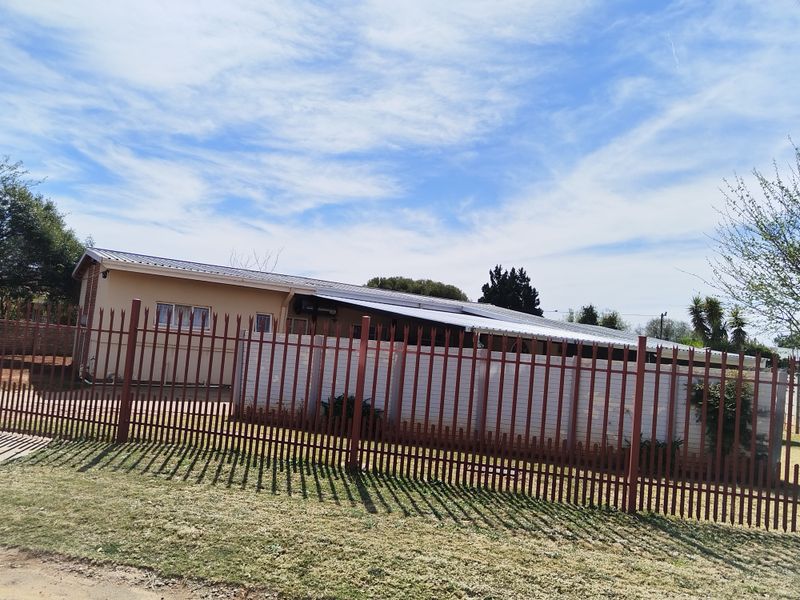 A view of the property's front exterior, featuring plastered walls, a corrugated metal roof, and secure metal perimeter fencing.