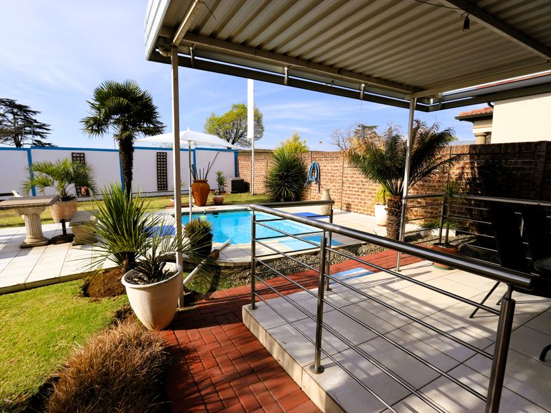 Outdoor entertainment area featuring a covered patio with tiled flooring, metal railings, and a swimming pool set within a landscaped garden.
