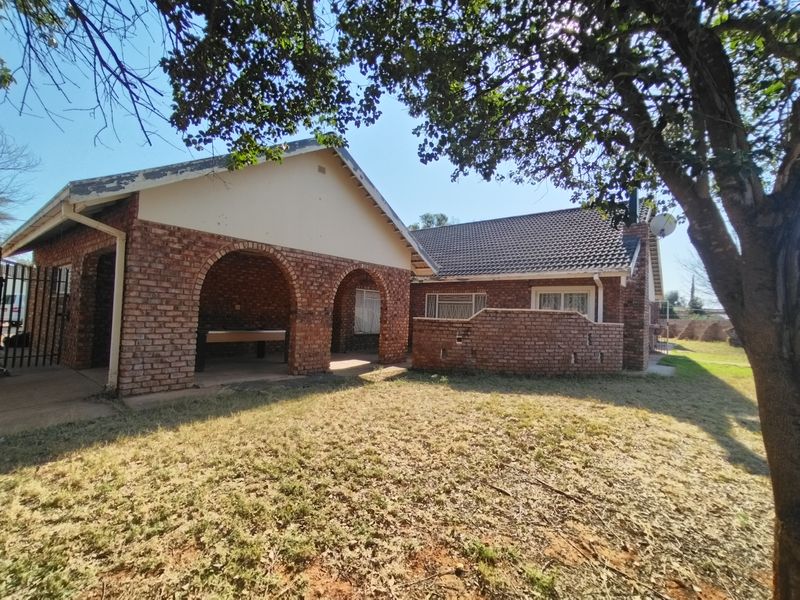 Rear view of a brick house featuring an arched patio area and a tiled roof, set within a garden with mature trees.