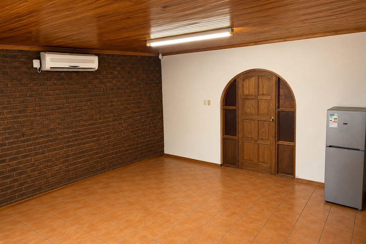 An empty living area featuring a timber panelled ceiling, exposed brick feature wall, ceramic floor tiles, and a distinctive arched timber entrance door.