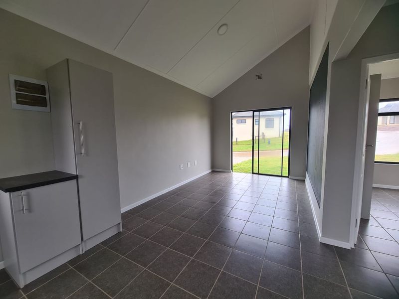 A bright, open-plan living space featuring vaulted ceilings, neutral grey walls, dark tiled flooring, and a sliding glass door leading to the exterior.