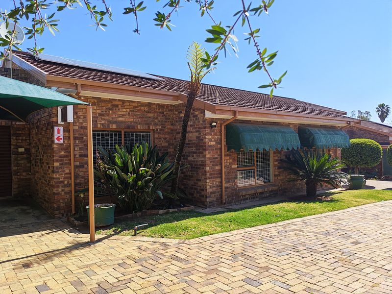 A well-maintained face brick townhouse exterior featuring a tiled roof, solar panels, paved driveway, and secure window treatments.