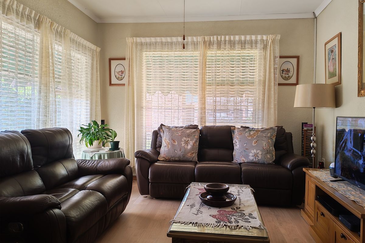 A comfortable living room featuring light-toned laminate flooring, coved ceilings, and large windows fitted with venetian blinds and sheer curtains.