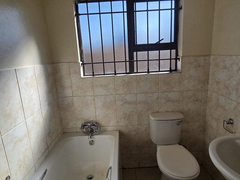 A functional bathroom featuring a bathtub, pedestal basin, and toilet, finished with neutral wall tiles and security bars on the window.