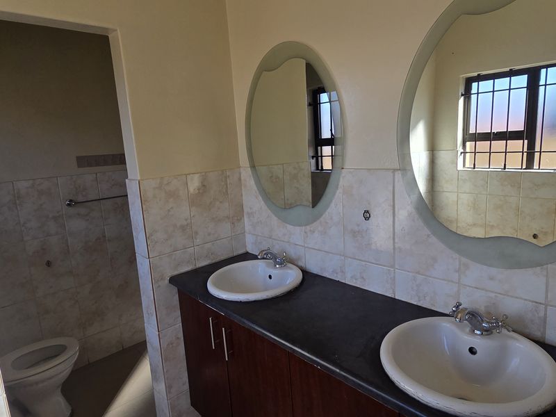 A functional bathroom featuring a double vanity with ceramic basins, tiled splashbacks, and a separate toilet area with security bars on the window.
