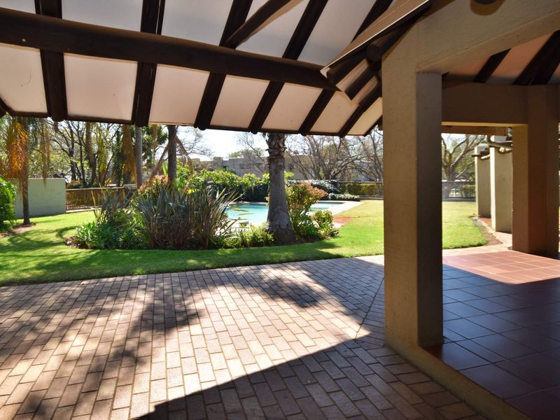 A spacious covered patio featuring exposed timber beams and paved flooring, overlooking a private swimming pool and well-maintained garden area.