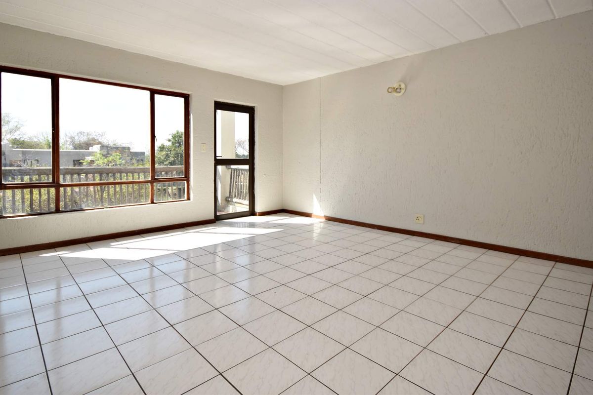 A bright, empty living room featuring white tiled flooring, textured walls, and large windows providing natural light and access to an outdoor balcony area.