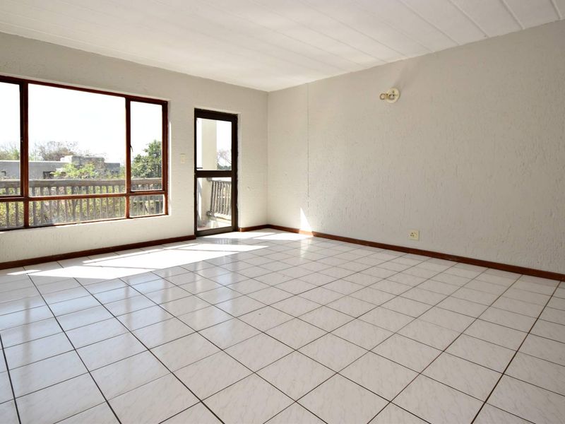 A bright, empty living room featuring white tiled flooring, textured walls, and large windows providing natural light and access to an outdoor balcony area.