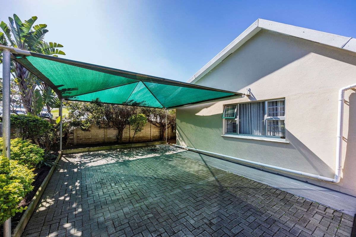 Rear exterior view featuring a paved parking area with a green shade cloth carport structure and a precast concrete boundary wall.