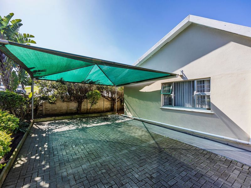 Rear exterior view featuring a paved parking area with a green shade cloth carport structure and a precast concrete boundary wall.