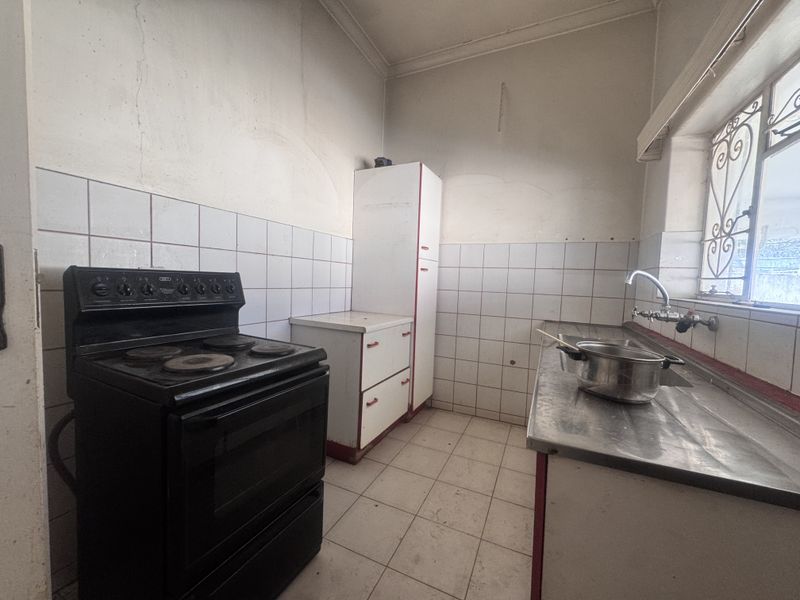 A dated kitchen featuring a freestanding electric stove, stainless steel sink, tiled backsplash, and original cabinetry in need of renovation.