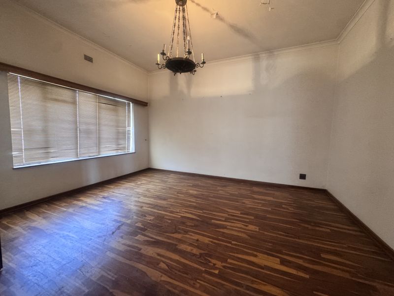 An empty living room featuring hardwood flooring, a classic chandelier, and large windows with venetian blinds, requiring minor cosmetic updates to the ceiling and walls.
