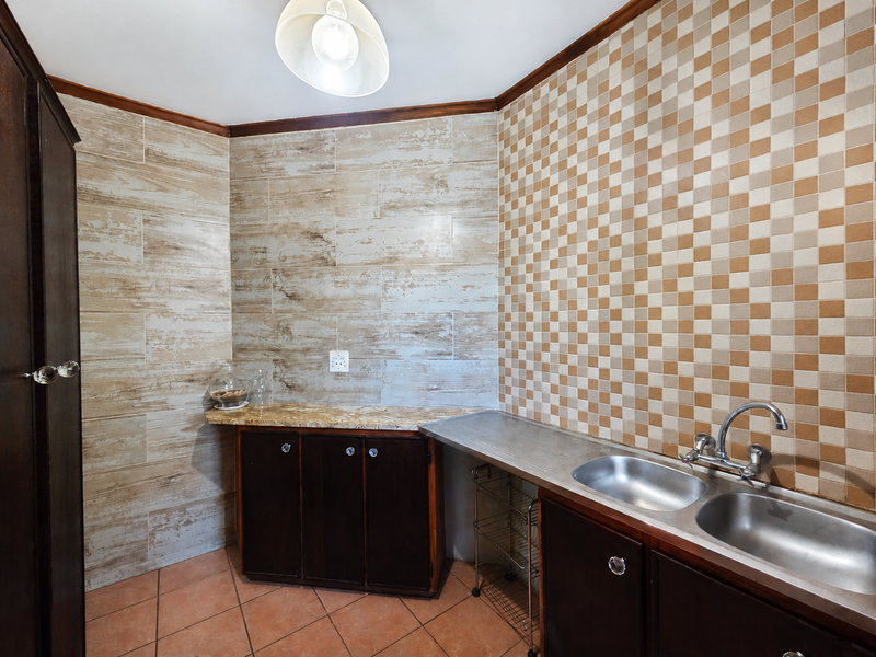 A functional scullery area featuring double stainless steel sinks, built-in dark cabinetry, stone-look countertops, and decorative tiled wall finishes.