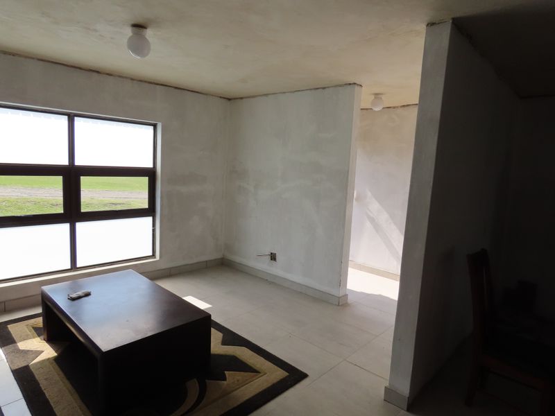 An empty living room space featuring tiled flooring, large aluminium windows, and unfinished plaster walls, offering a blank canvas for renovation.