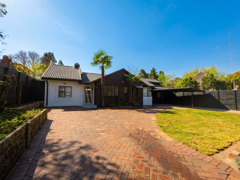 A well-maintained front exterior featuring a spacious brick-paved driveway, a carport, and secure perimeter fencing with established landscaping.