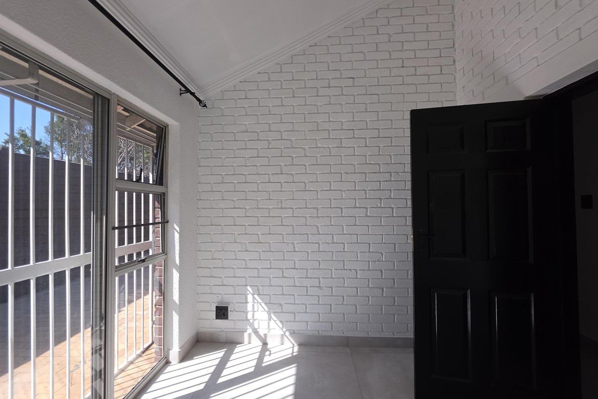 A bright, empty bedroom featuring a white painted brick feature wall, vaulted ceiling, tiled floors, and a sliding glass door with security bars.