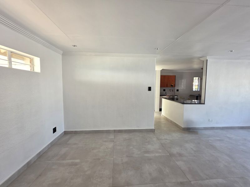 An open-plan living area featuring large format floor tiles, recessed lighting, and a view through to the kitchen with built-in cabinetry.