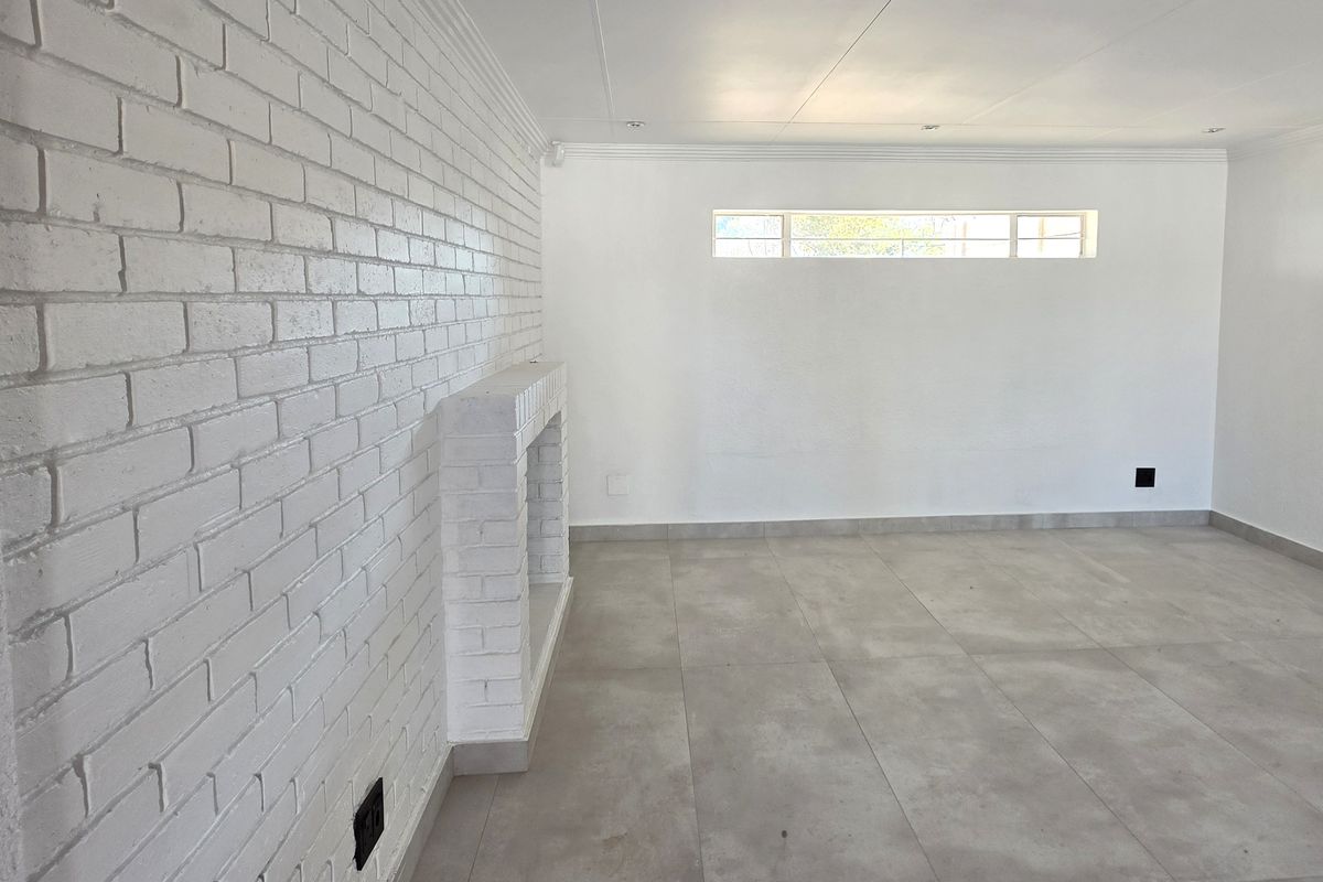 A bright, empty living room featuring a white painted brick wall with a built-in fireplace, modern grey floor tiles, and recessed ceiling lighting.