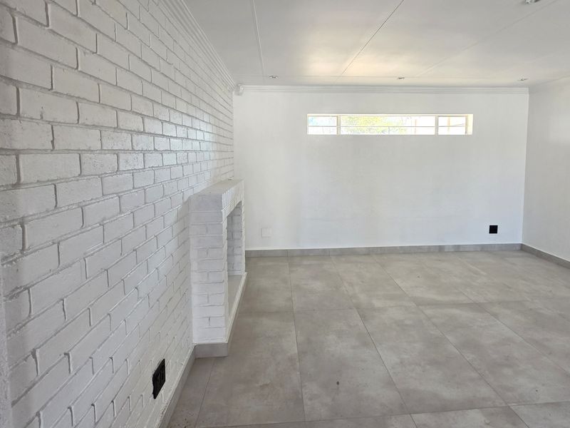 A bright, empty living room featuring a white painted brick wall with a built-in fireplace, modern grey floor tiles, and recessed ceiling lighting.