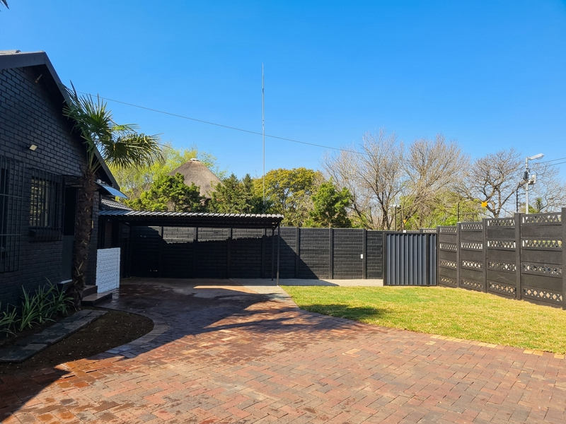 A well-maintained rear exterior area featuring a brick-paved driveway, a covered carport, secure perimeter fencing, and a neat lawn.