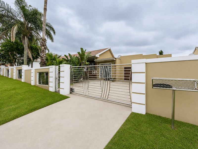 A well-maintained front exterior featuring a modern stainless steel driveway gate, perimeter boundary wall, and a neatly landscaped lawn.