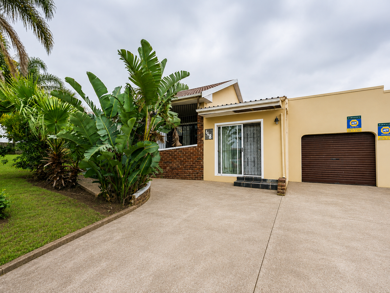 Front view of the property featuring a concrete driveway, double garage with roller doors, and a landscaped garden area with mature plants.