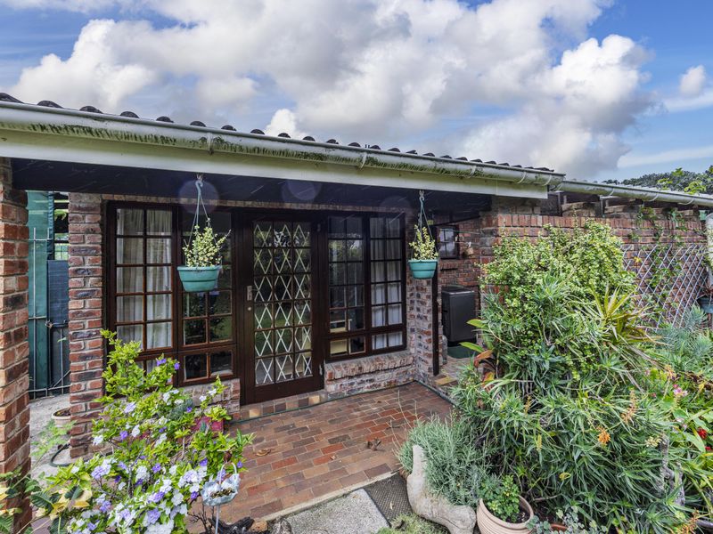 A covered brick patio area featuring a timber-framed glass entrance door and windows, overlooking a landscaped garden space.