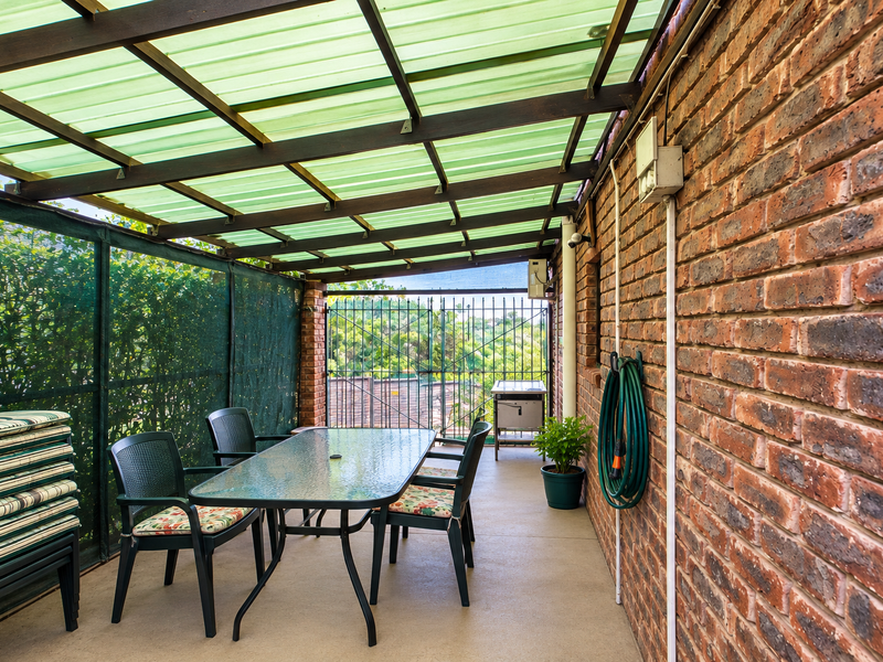 A covered patio area featuring an exposed brick wall, timber roof structure with translucent sheeting, and a security gate overlooking the garden.