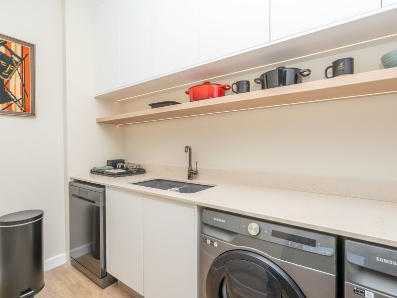 Modern laundry area featuring sleek white cabinetry, stone benchtops, an undermount sink, integrated dishwasher, and stylish under-shelf lighting.