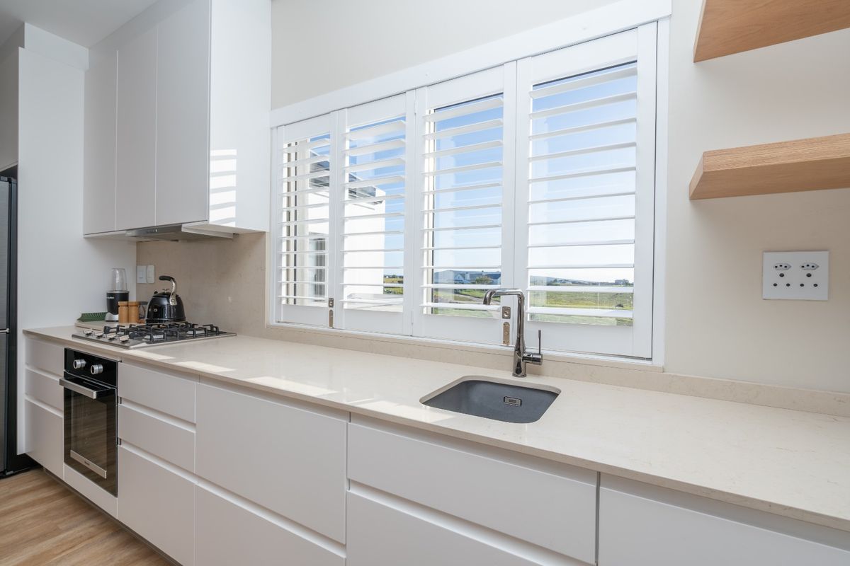 Modern kitchen featuring sleek white cabinetry, stone countertops, integrated gas hob, oven, and stylish plantation shutters overlooking the exterior.