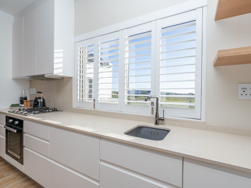 Modern kitchen featuring sleek white cabinetry, stone countertops, integrated gas hob, oven, and stylish plantation shutters overlooking the exterior.