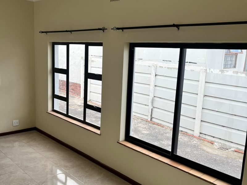 A bright, empty living area featuring neutral tiled flooring, modern black aluminium window frames, and clean wall finishes.