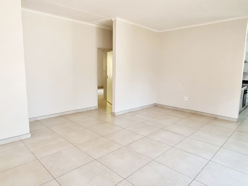 A bright, open-plan living and kitchen area featuring neutral tiled flooring, clean white walls, and a solid wood door leading to adjacent rooms.