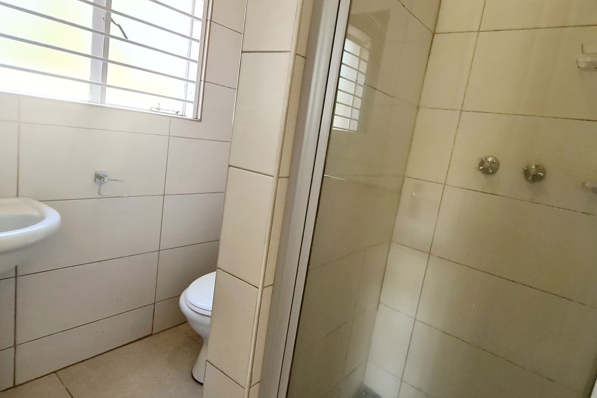A functional bathroom featuring a pedestal basin, toilet, and a glass-enclosed shower with tiled walls and security bars on the window.