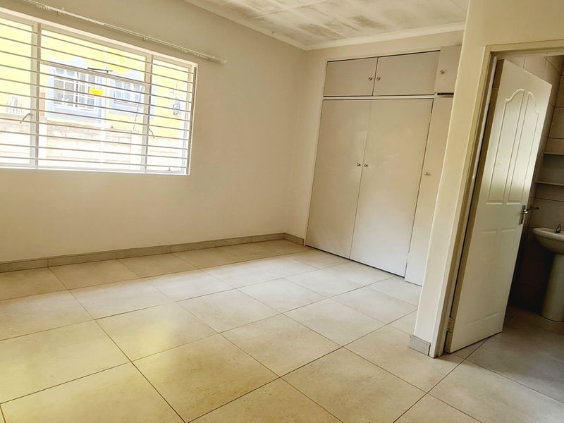 An empty bedroom featuring tiled flooring, built-in cupboards, and an adjoining en-suite bathroom with a pedestal basin.