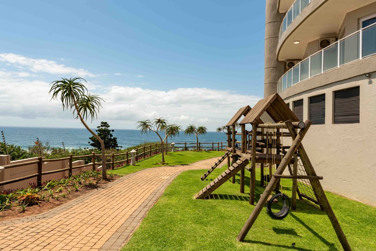 A well-maintained rear garden area featuring a paved walkway, lawn, and ocean views, adjacent to the building's exterior facade and balcony.