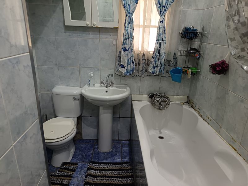 A compact bathroom featuring a pedestal basin, toilet, and bathtub with fully tiled walls and flooring, requiring some maintenance to finishes.