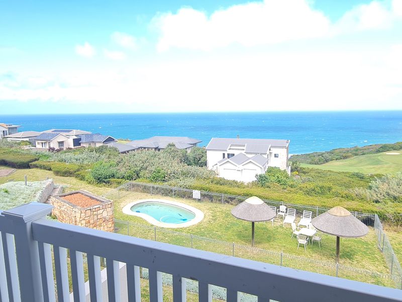 Elevated coastal view overlooking a residential swimming pool area, landscaped grounds, and the ocean horizon.