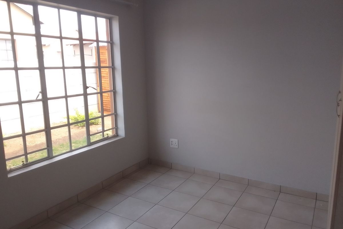 A bright, empty bedroom featuring neutral tiled flooring, a large window with security bars, and a clean, minimalist aesthetic.