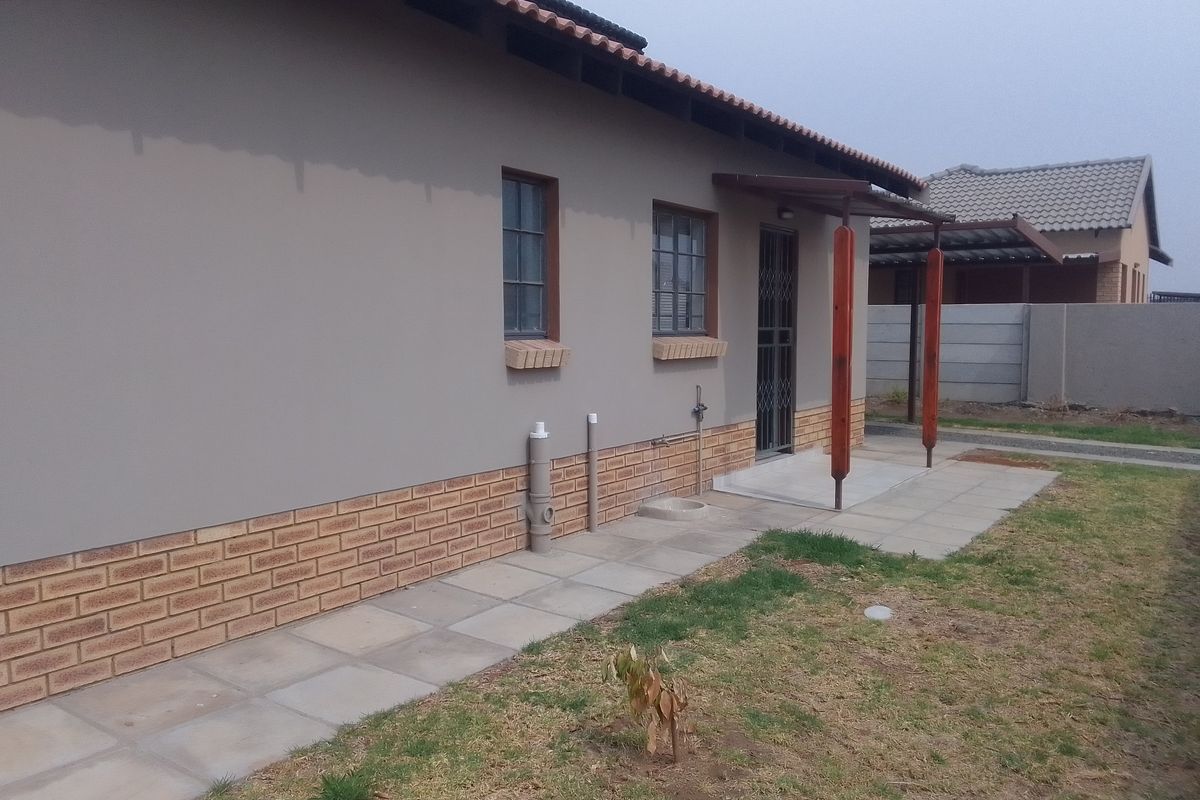 Rear exterior view of the townhouse featuring a paved walkway, covered patio area, and brick-accented facade with security gate.